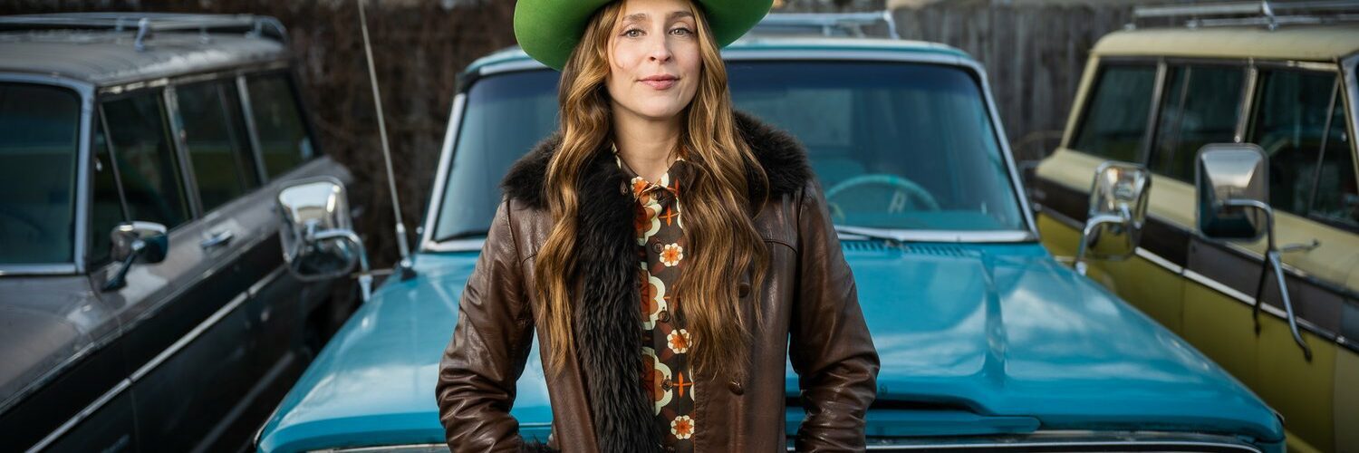Singer Angela Perley in a green hat standing in front of a blue car