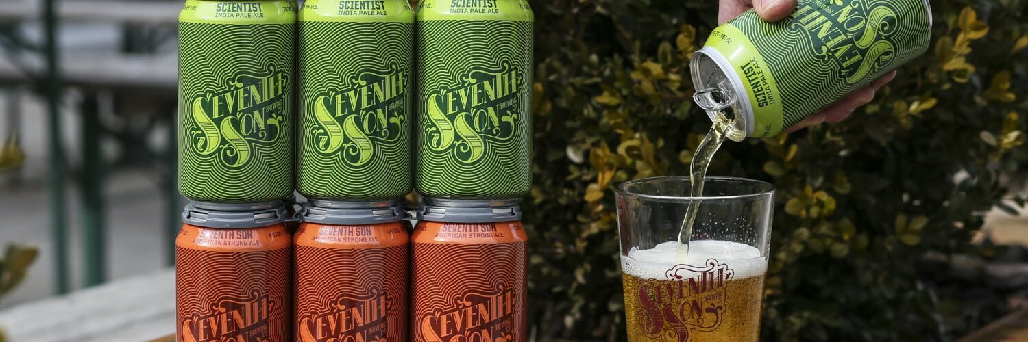 A yellow six pack of Seventh Son beer stacked on top of an orange six pack, next to a hand pouring beer into a pint glass.